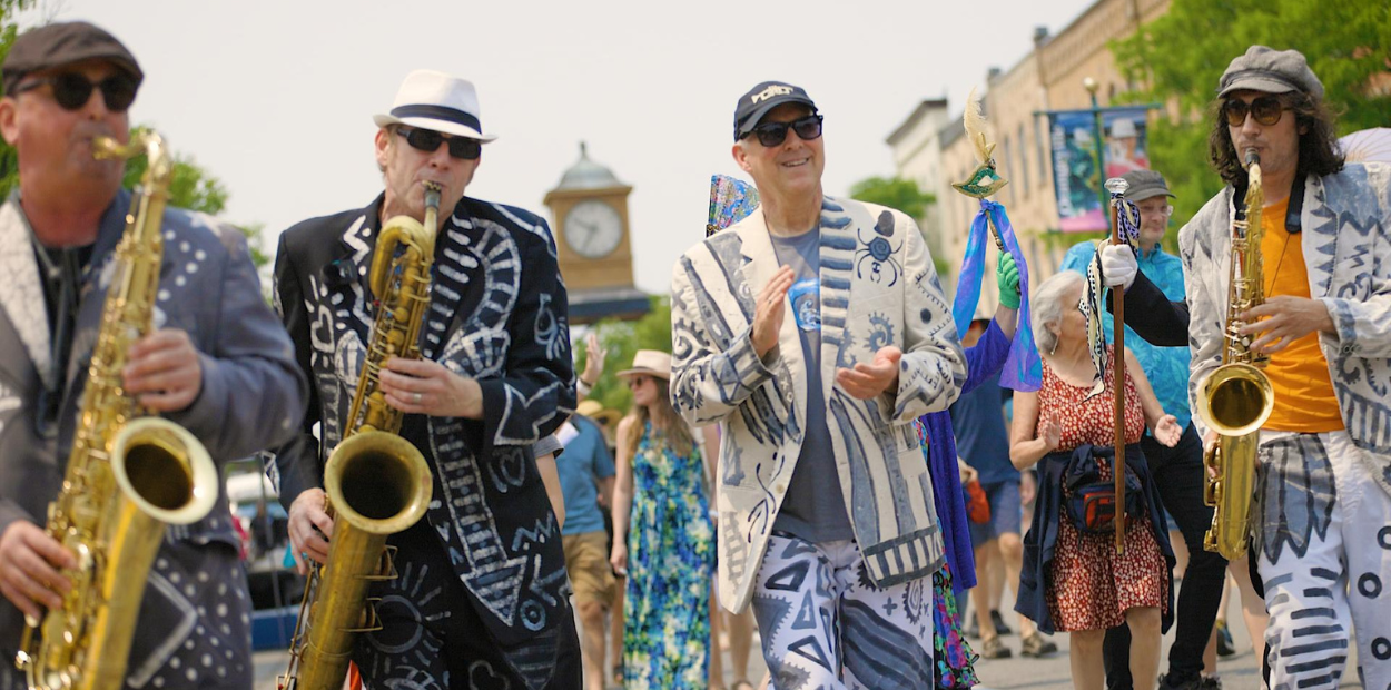 Three men with saxophones and two men clapping while marching in a parade.